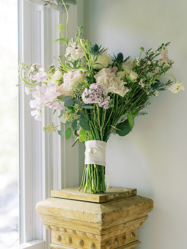 A bouquet of pastel flowers and greenery wrapped with white ribbon and a pearl pin rests on a decorative wooden pedestal near a bright window.