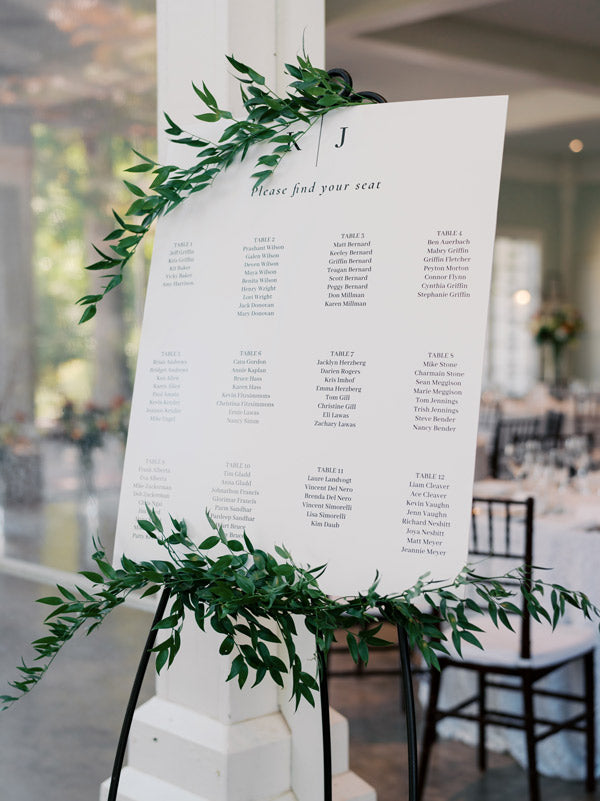 A wedding seating chart decorated with green leafy garlands is displayed on an easel. The chart lists guests’ names organized by table under the heading “Please find your seat” in an elegant, indoor venue.