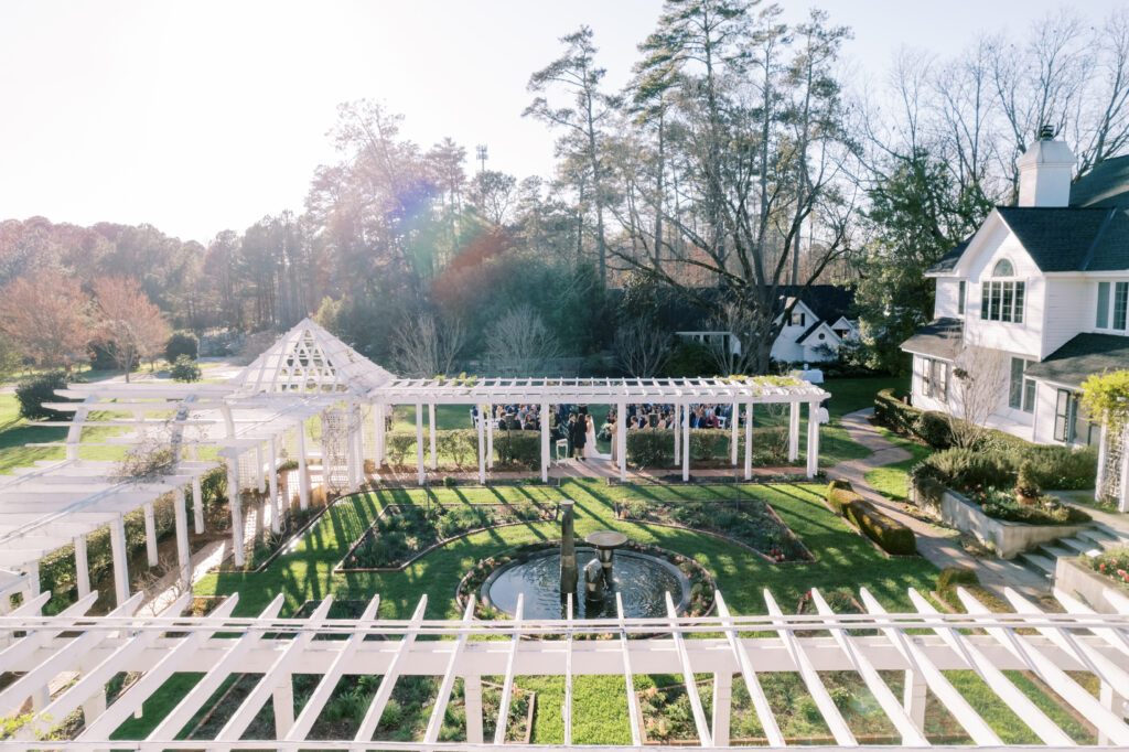 Aerial view of a white pergola and fountain in a landscaped garden, surrounded by tall trees and a large white house on a sunny day.