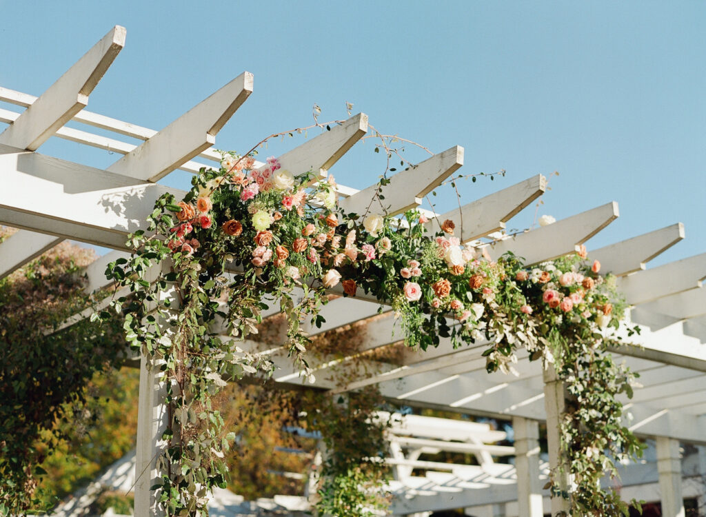 A white pergola is decorated with lush greenery and clusters of pink, peach, and orange flowers under a clear blue sky.