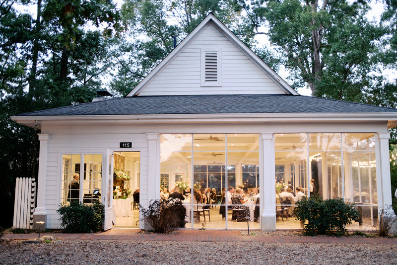 A white, single-story building with large windows shows people gathered inside for an event, possibly a dinner or reception, surrounded by trees and lit warmly from within during the evening.