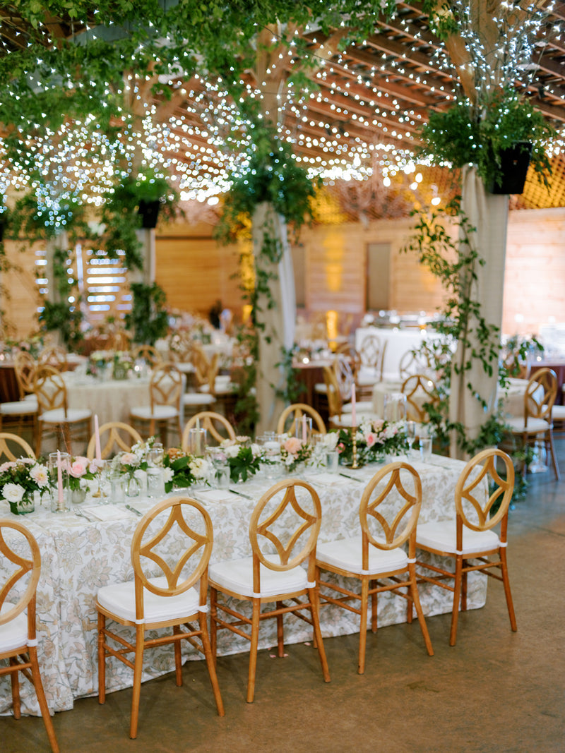 A decorated indoor wedding reception with round and rectangular tables covered in floral-patterned cloths, wooden chairs, lush greenery, and twinkling string lights hanging from the ceiling.