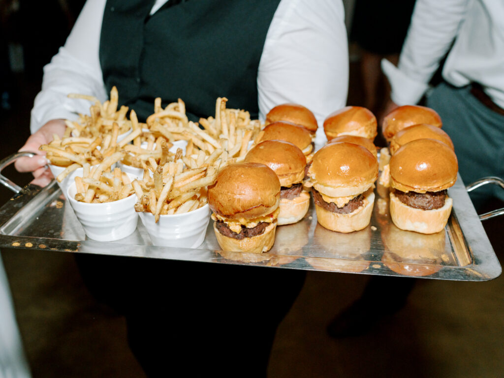 A server holding a metal tray with six mini cheeseburgers and three white cups filled with French fries, offering them at an event.