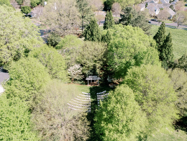 Aerial view of a green, tree-filled outdoor area with rows of white chairs arranged in front of a wooden arbor, likely set up for a ceremony, surrounded by residential houses and open grassy spaces.