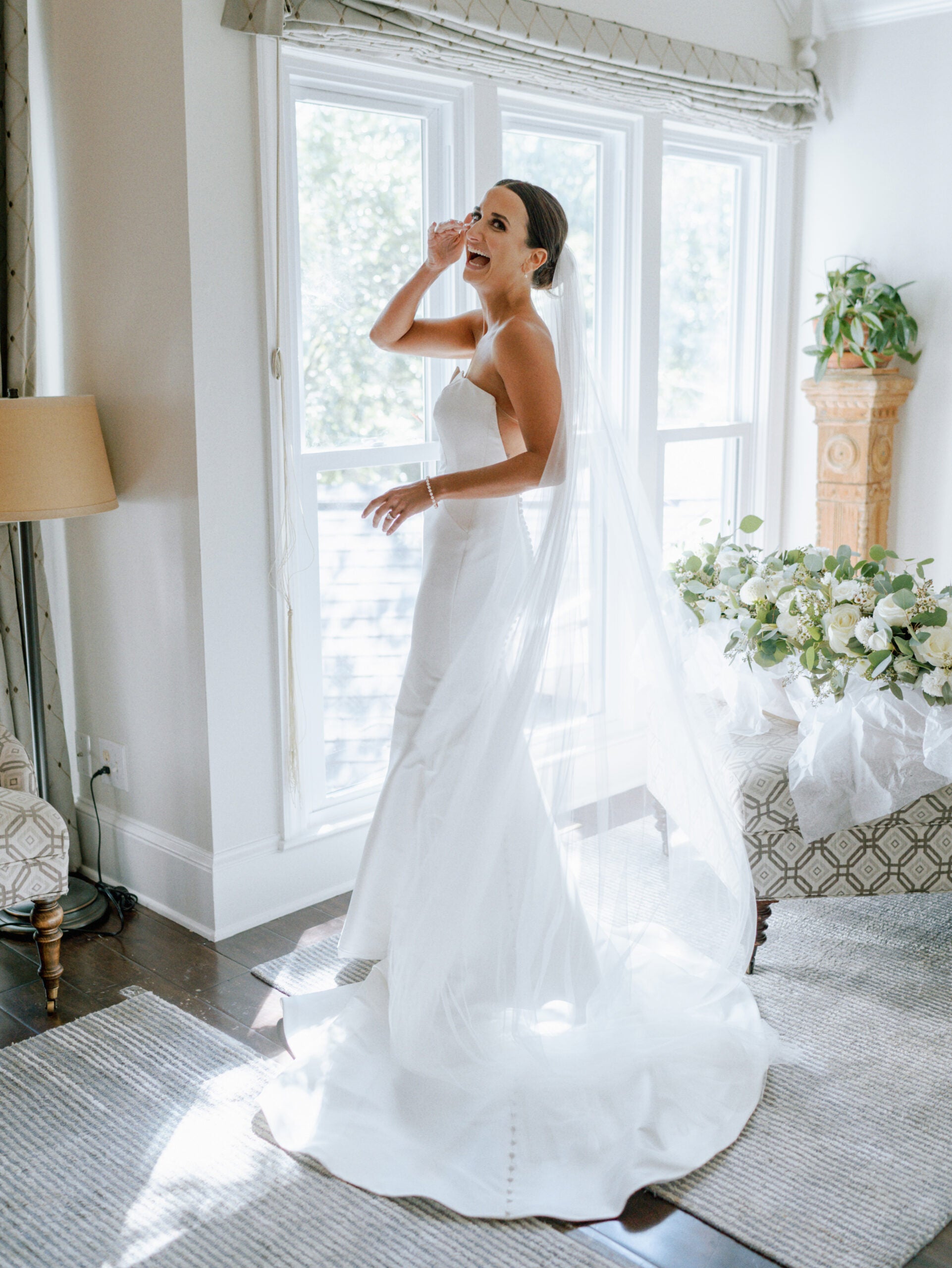 A bride in a strapless white gown and long veil stands smiling near large windows, holding a tissue to her face. Sunlight fills the elegant room decorated with white flowers and neutral furnishings.