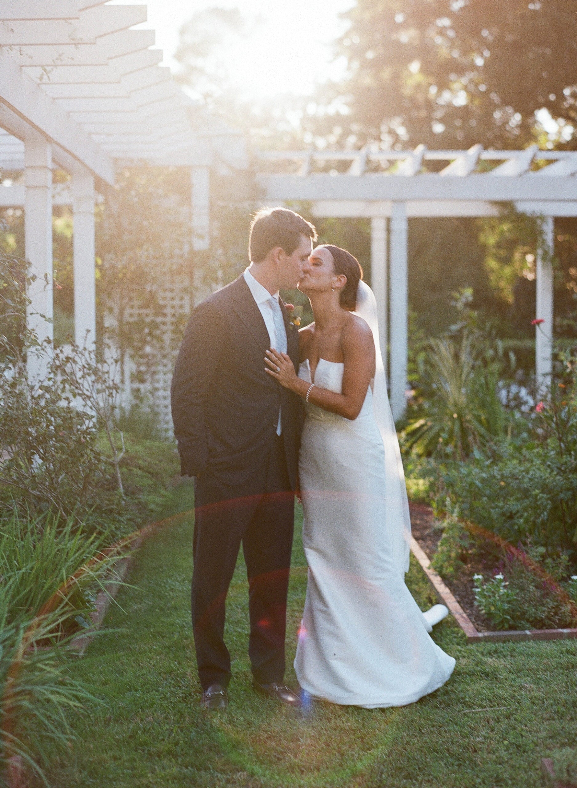 A bride in a strapless white gown kisses a groom in a dark suit on the cheek as they stand in a sunlit garden with greenery and a white pergola in the background.