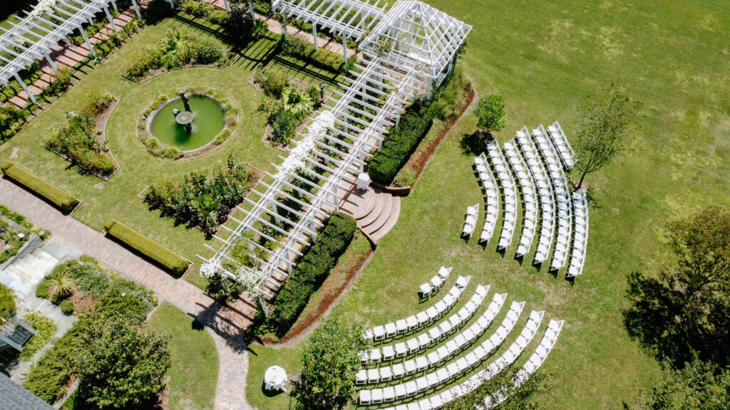 Aerial view of an outdoor wedding venue with white chairs arranged in rows on a lawn, a circular fountain, brick paths, greenery, and a white pergola structure.