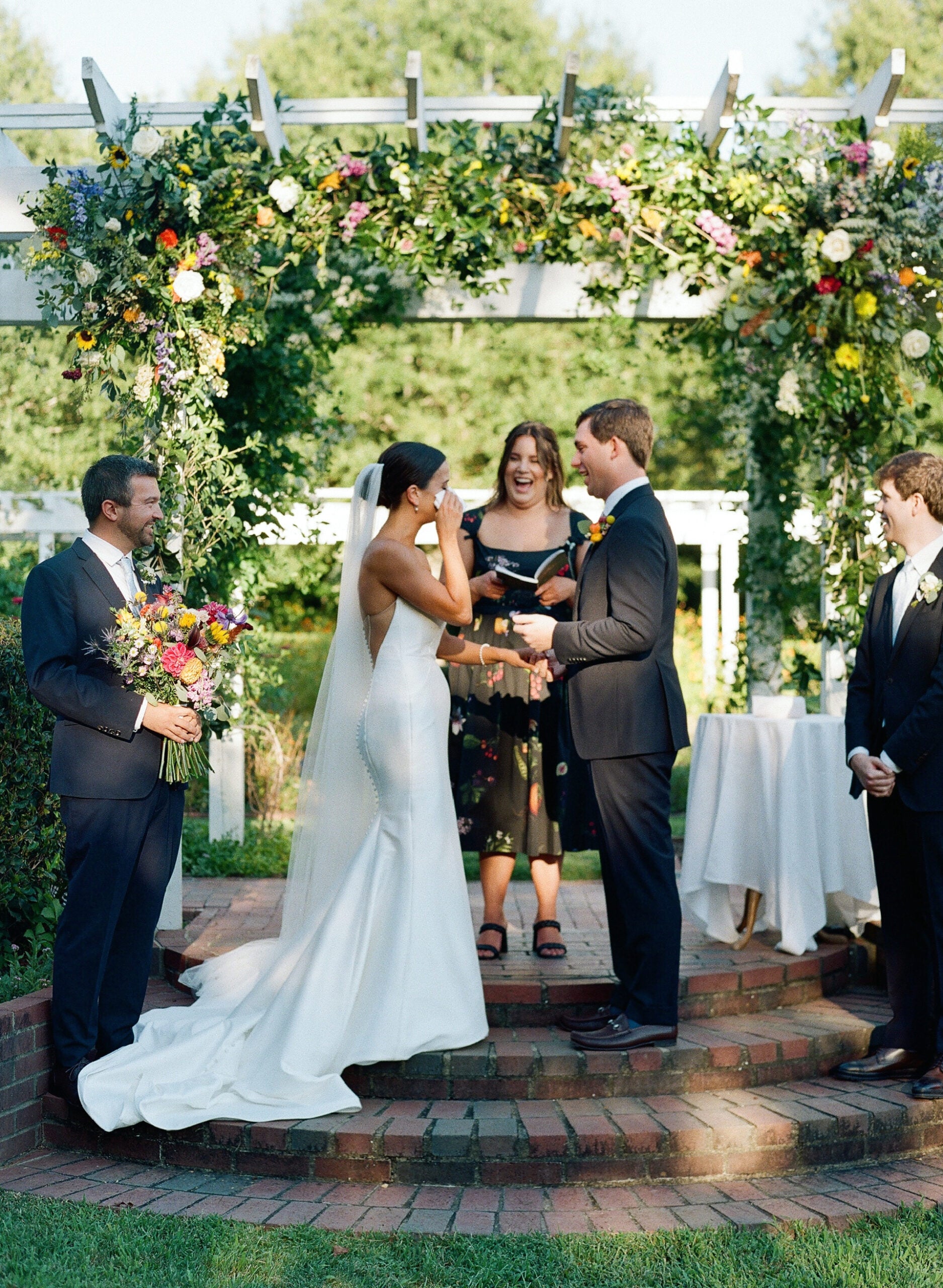 A bride wipes away tears while standing with her groom during an outdoor wedding ceremony under a lush floral arch, surrounded by the officiant and two groomsmen.