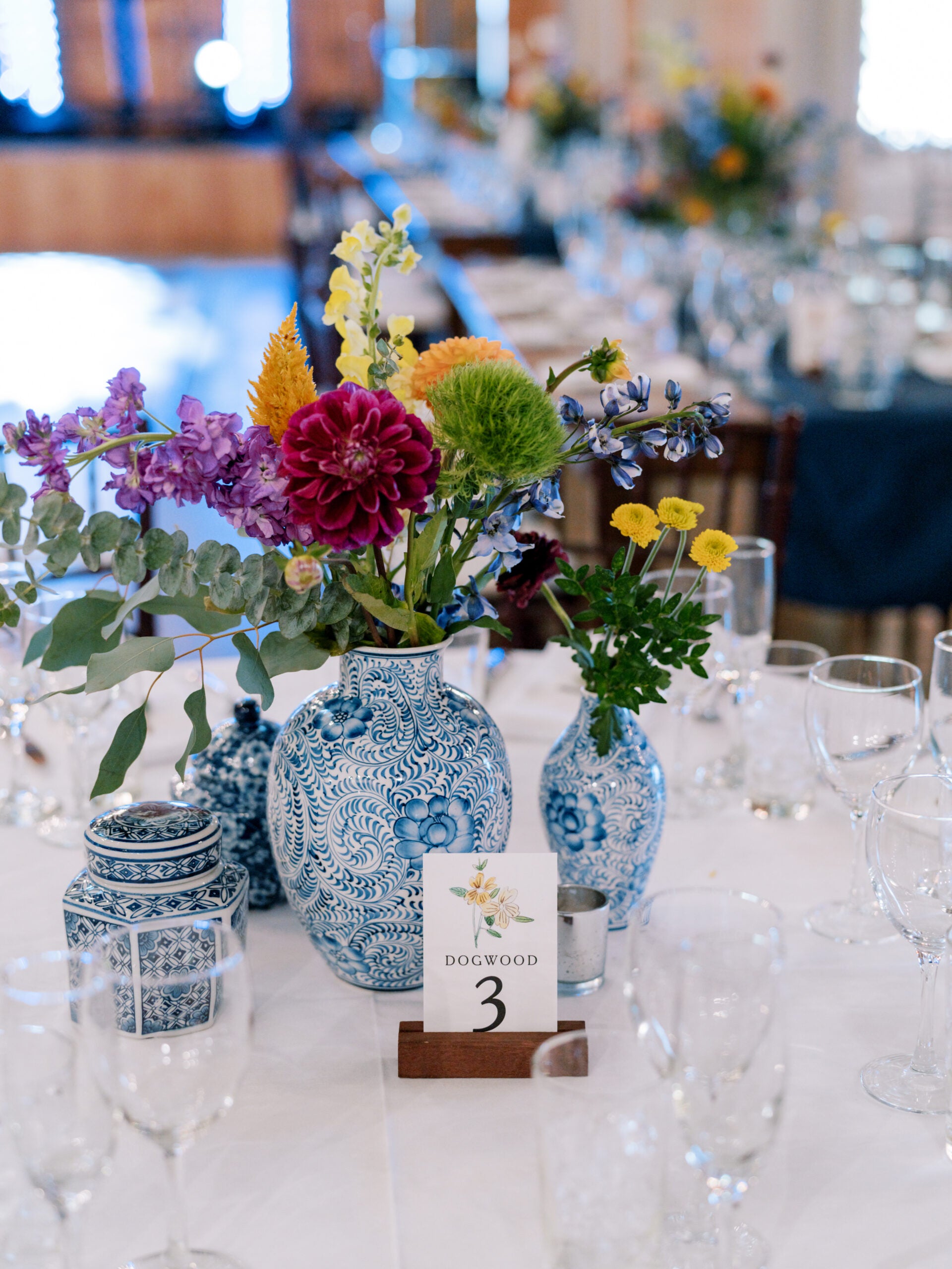 A table centerpiece featuring colorful flowers in blue and white patterned vases, surrounded by clear wine glasses, with a card labeled 