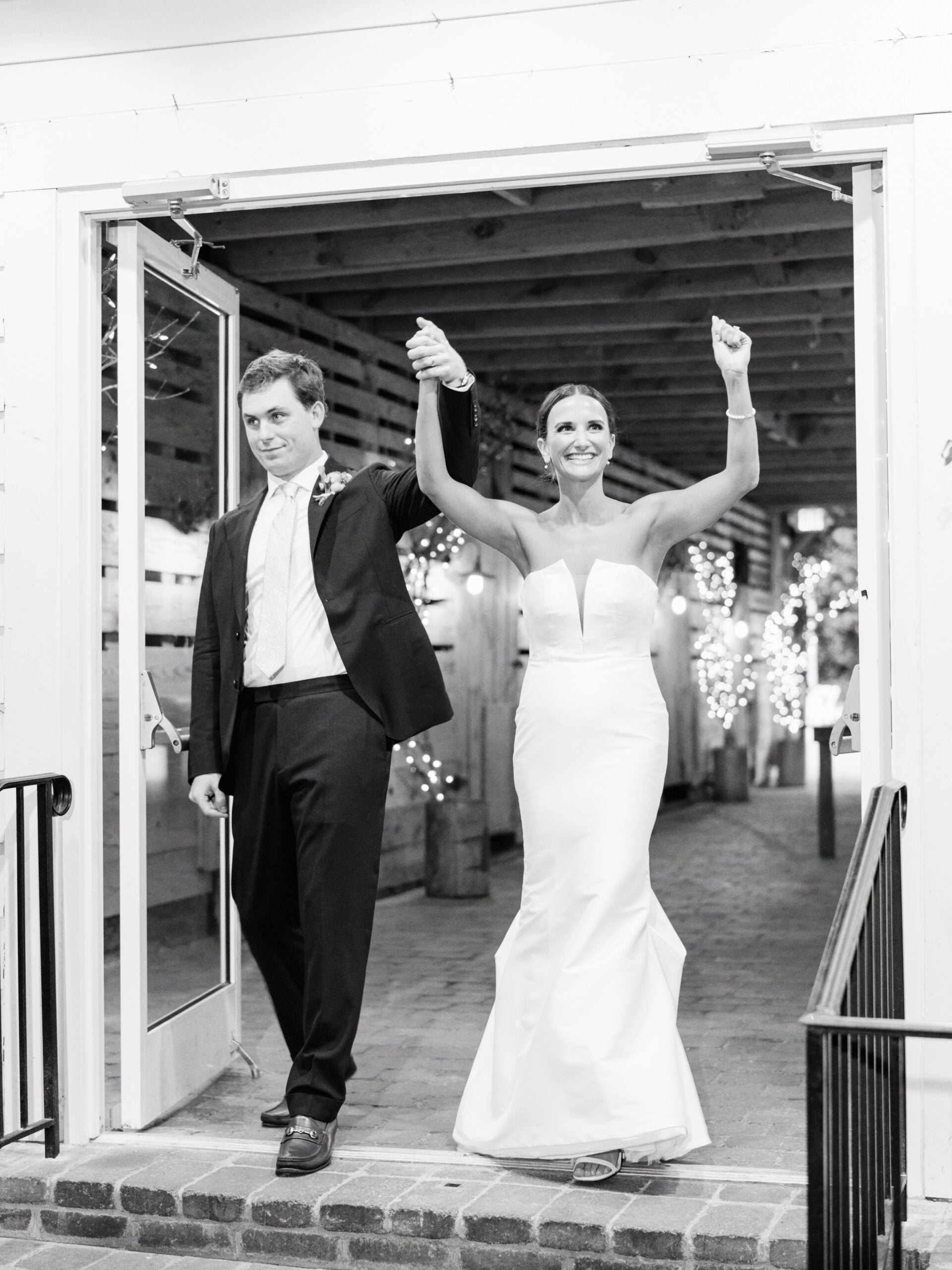 A bride and groom celebrate as they enter a venue, holding hands with arms raised. The bride wears a strapless white gown and smiles, while the groom in a suit walks beside her. String lights decorate the background.