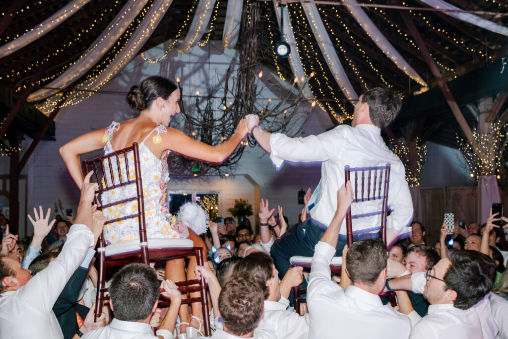 A bride and groom are lifted on chairs by guests during a lively wedding celebration, surrounded by cheering people under strings of lights and draped fabric in a decorated venue.