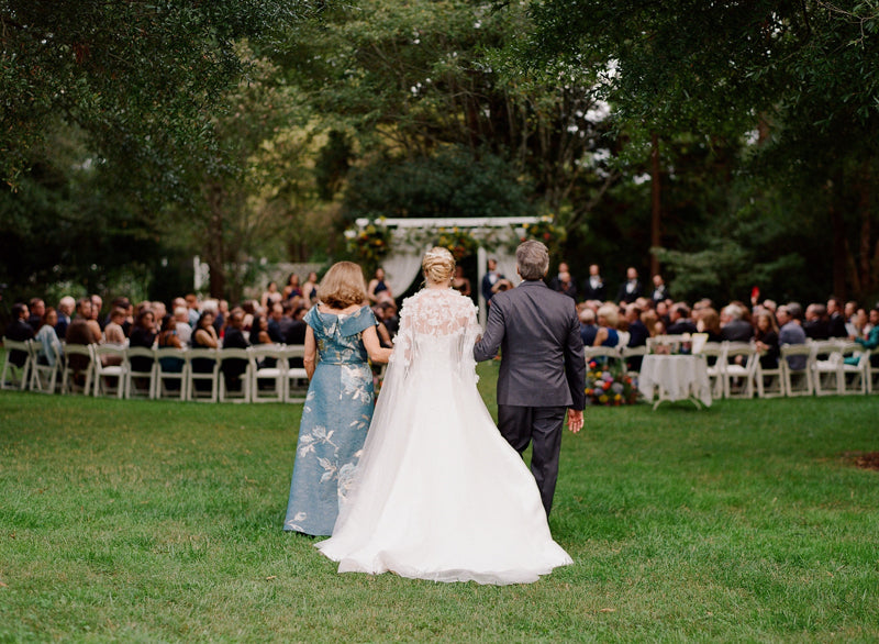 A bride in a white gown walks between two adults down a grassy aisle toward a seated crowd at an outdoor wedding ceremony, with trees and a decorated archway in the background.