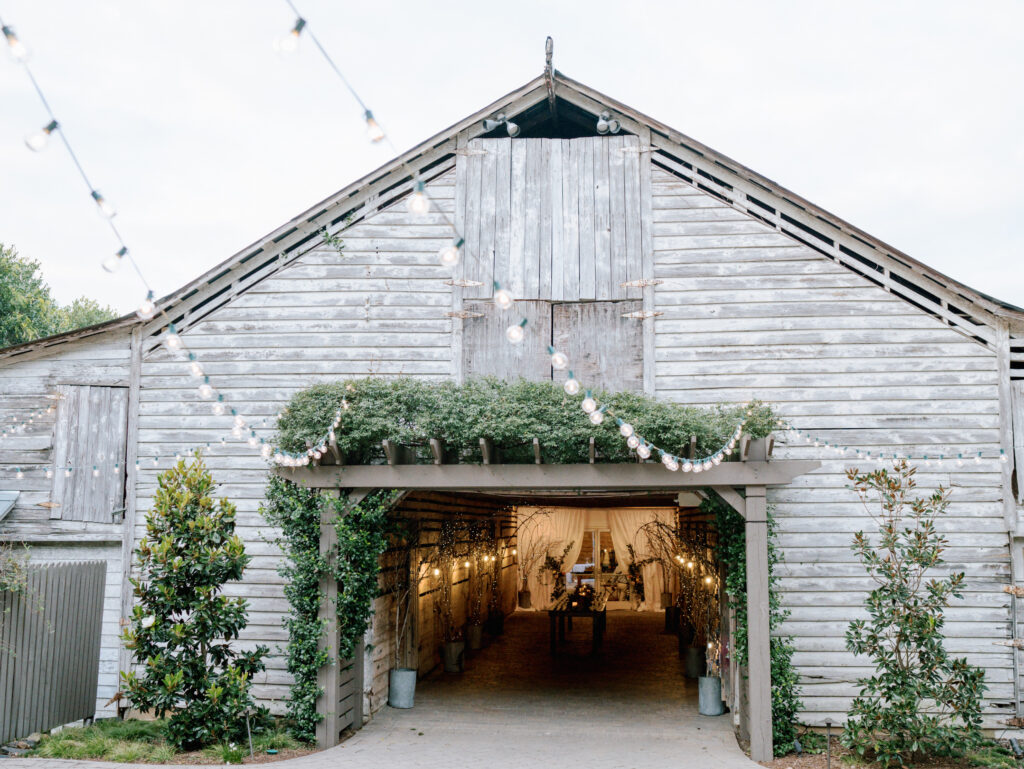 A rustic wooden barn decorated with string lights and greenery, with its large doors open to reveal a warmly lit interior set for an event or gathering.