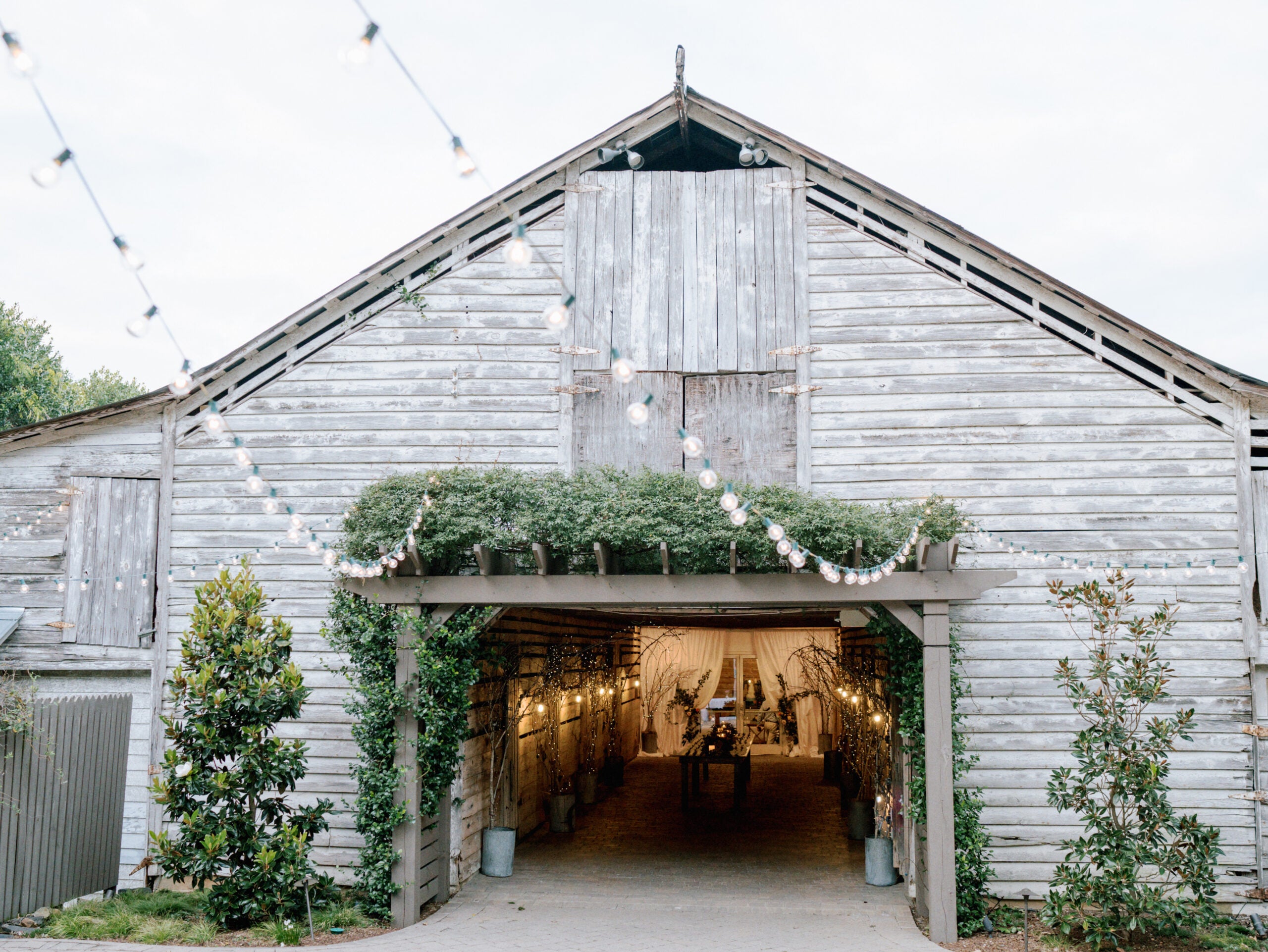 A rustic wooden barn decorated for an event, with string lights hanging outside and greenery framing the entrance; the interior is softly lit and visible through the open doorway.