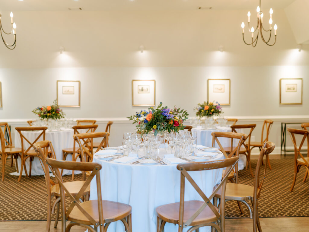 A banquet hall with round tables covered in white tablecloths, wooden chairs, elegant glassware, and floral centerpieces. Soft lighting and framed artwork decorate the cream-colored walls.