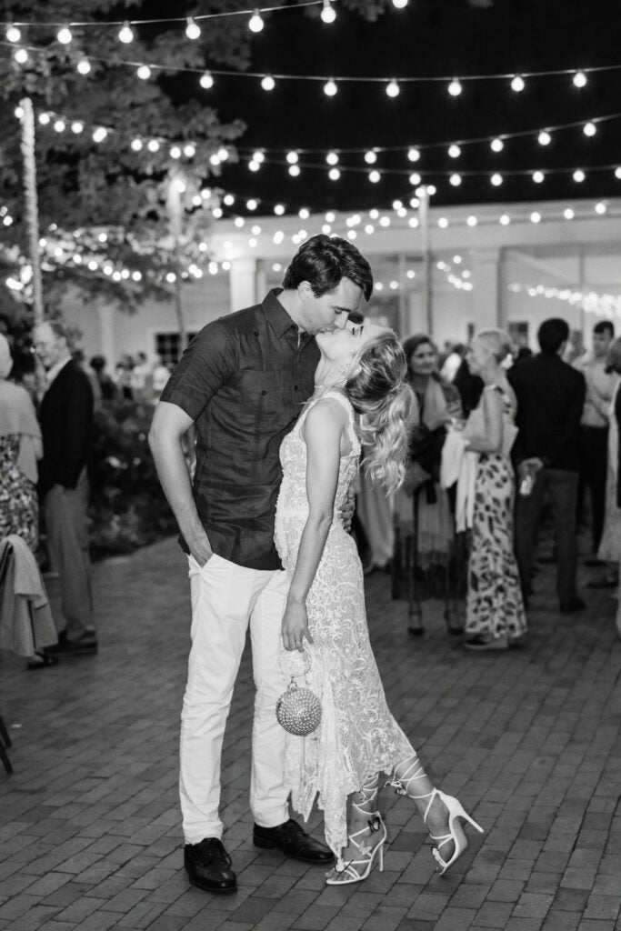 A couple kisses while standing close together at an outdoor evening event with string lights overhead. People in formal attire mingle in the background. The scene has a romantic, celebratory ambiance.