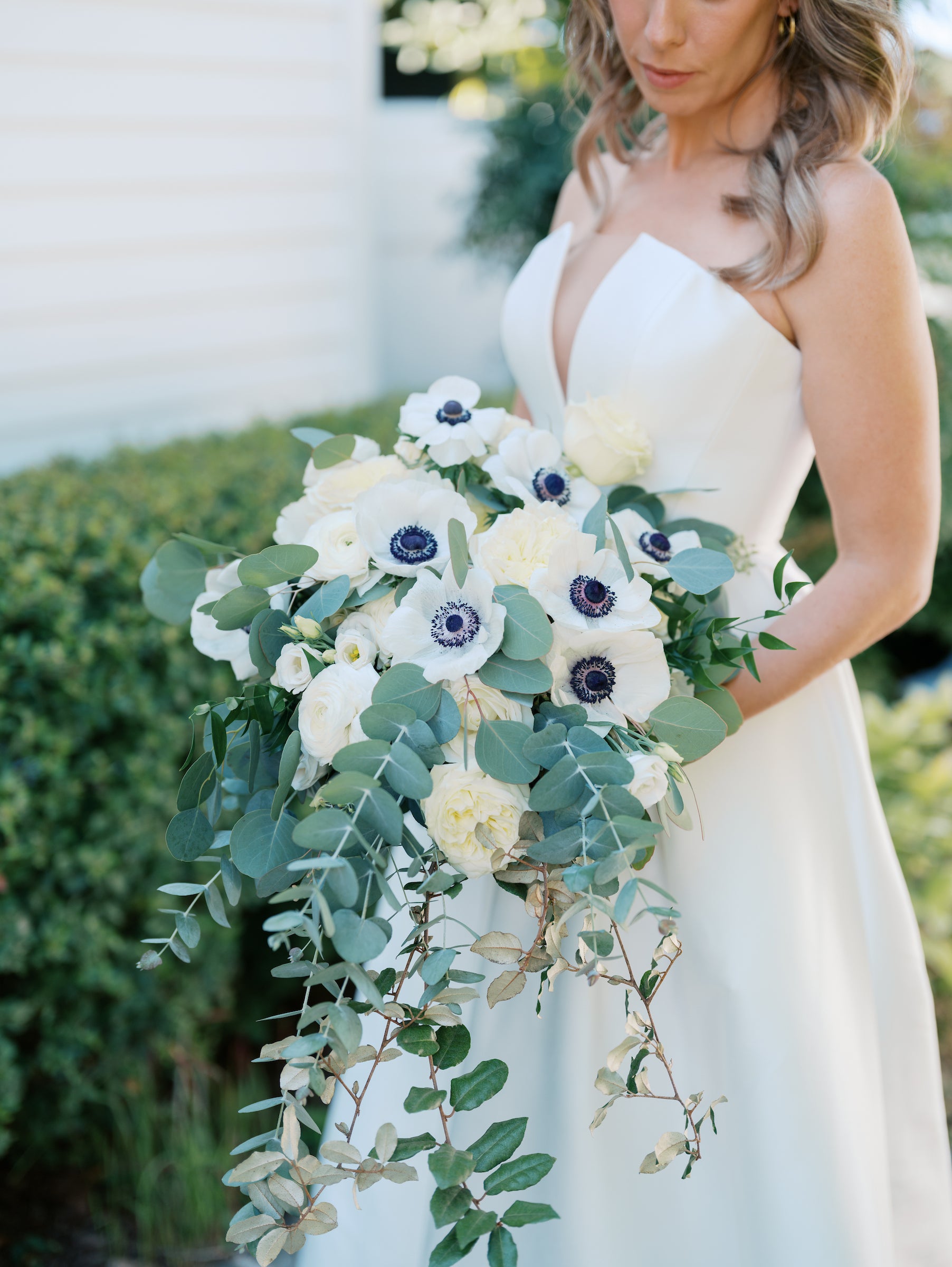 A bride in a white dress holds a large bouquet of white and purple flowers with green leafy foliage, standing outdoors near green bushes.