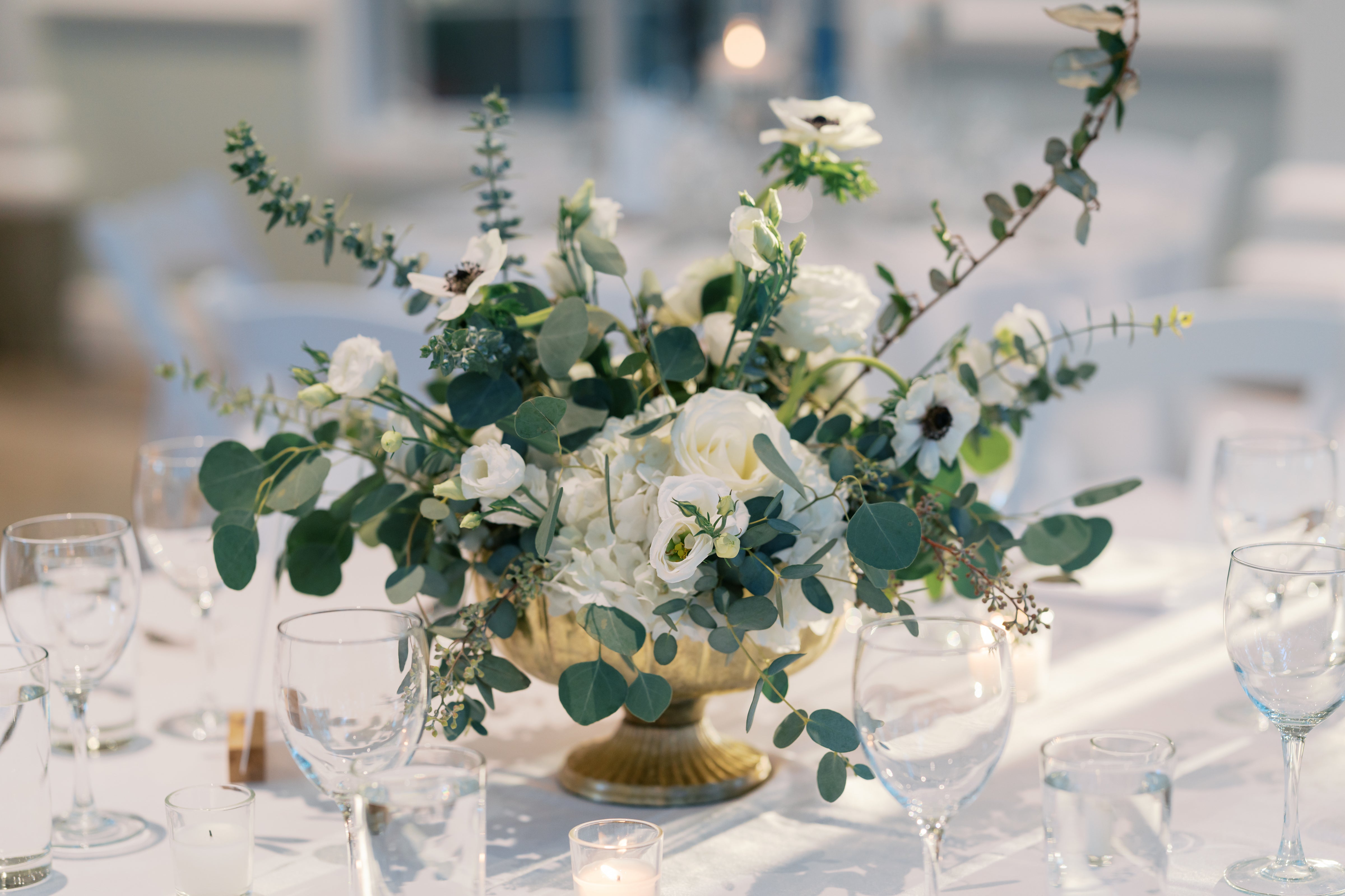 A gold vase filled with white flowers and greenery sits on an elegantly set table with clear wine glasses, small candles, and white tablecloths, creating a soft, romantic atmosphere.