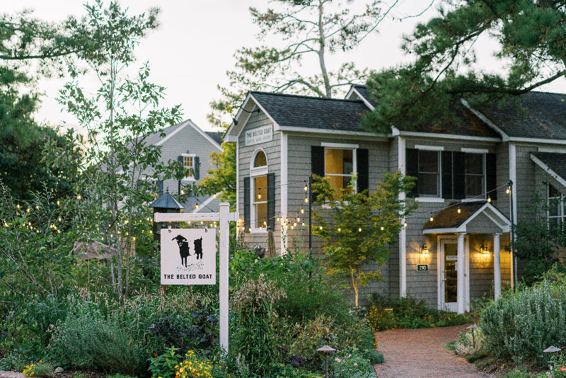 A gray, two-story cottage-style building surrounded by lush greenery and flowers. A white sign with a black goat logo and the words 