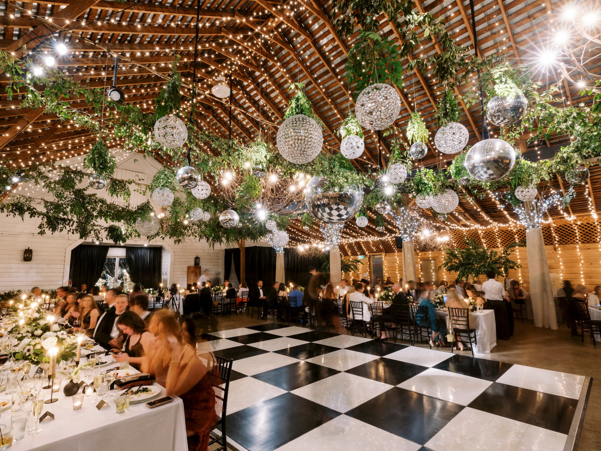 A decorated event hall with string lights, hanging greenery, and disco balls, featuring a black and white checkered dance floor. Guests sit at tables, dining and socializing under a wooden, vaulted ceiling.