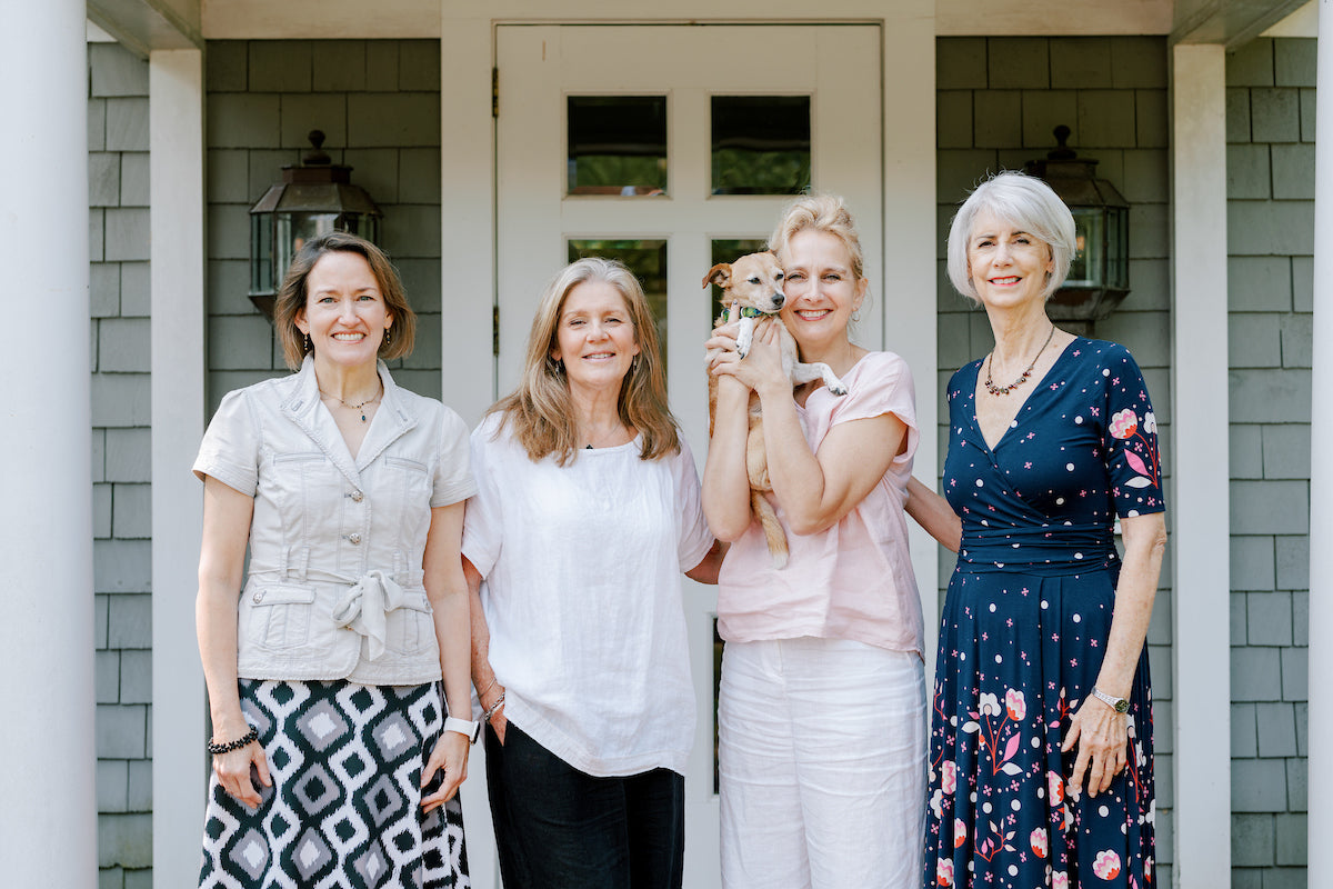 Four women stand together smiling in front of a house entrance; the third woman from the left holds a small dog. They are dressed in casual summer clothing.