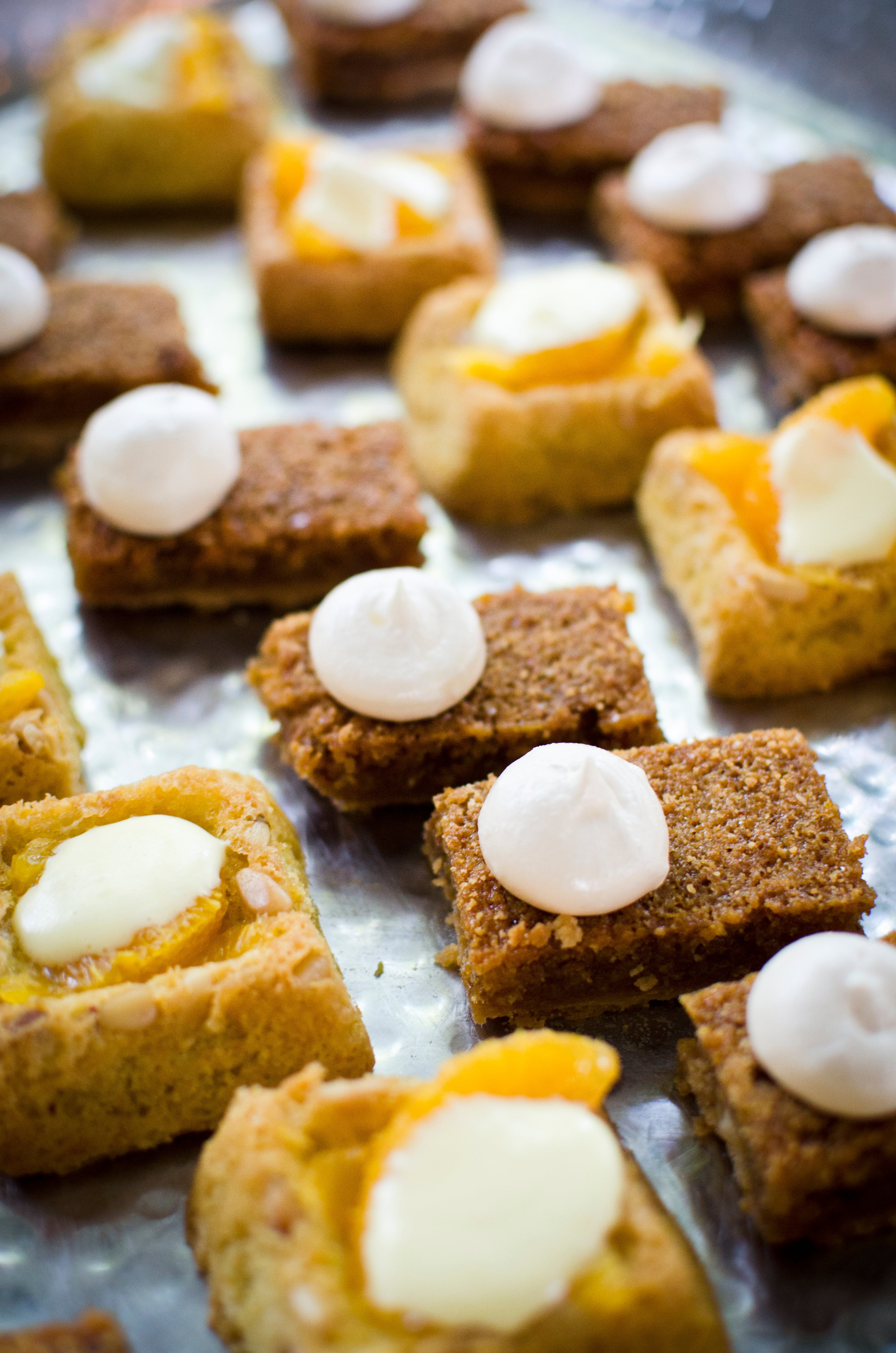Assorted bite-sized pastries on a tray, some topped with dollops of white cream and others with yellow fruit filling, arranged closely together on a metallic surface.