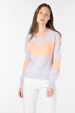 A person with long hair is wearing a KINROSS CASHMERE - GATHERED SLEEVE VEE NECK sweater in light purple with peach-colored accents, paired with white pants. They are standing in front of a plain white background, giving the look a comfortable yet stylish aesthetic.