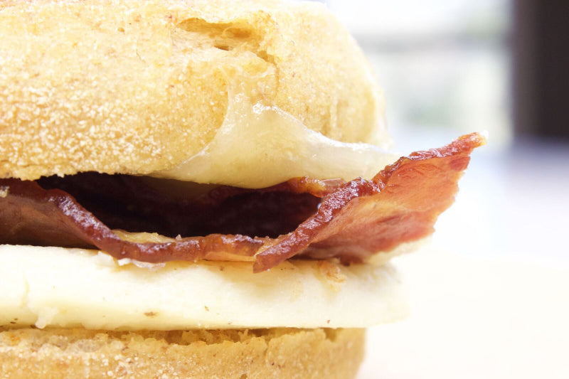 Close-up of a breakfast sandwich with crispy bacon, melted cheese, and egg, served on an English muffin. The image focuses on the sandwich's textures and layers.