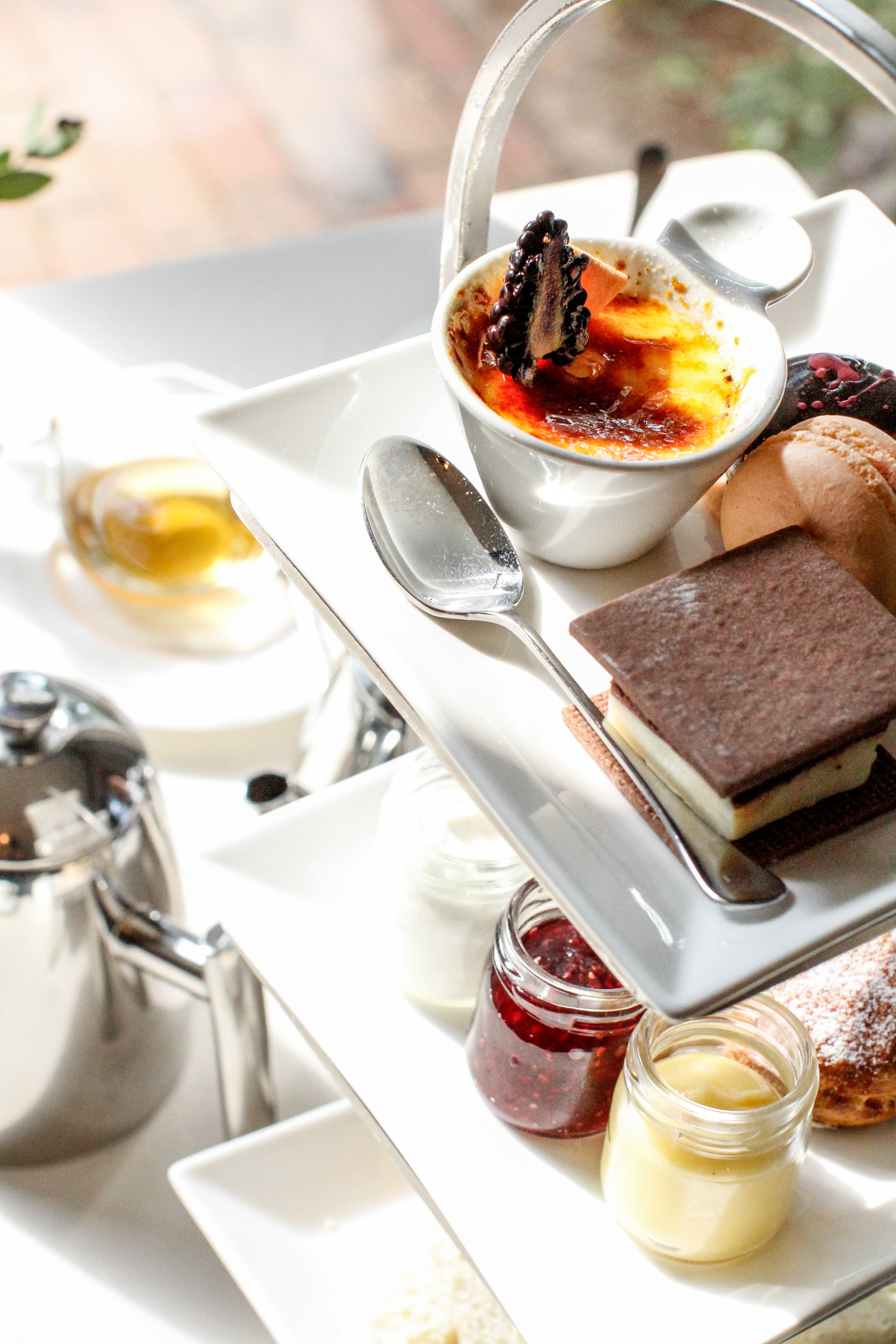 A close-up of a tiered tray with assorted desserts, including crème brûlée, layered cake, jams, and pastries, alongside a teapot and a cup of tea on a bright, sunlit table.