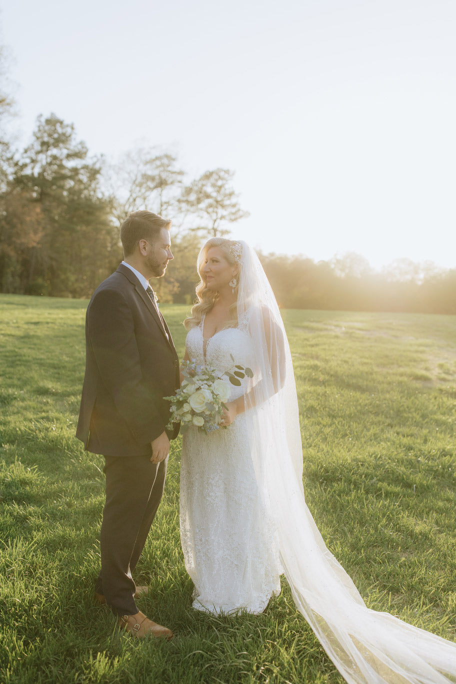 A bride and groom stand facing each other in a sunlit field. The bride wears a white lace gown and long veil, holding a bouquet, while the groom wears a dark suit. Trees and bright sunlight are visible in the background.