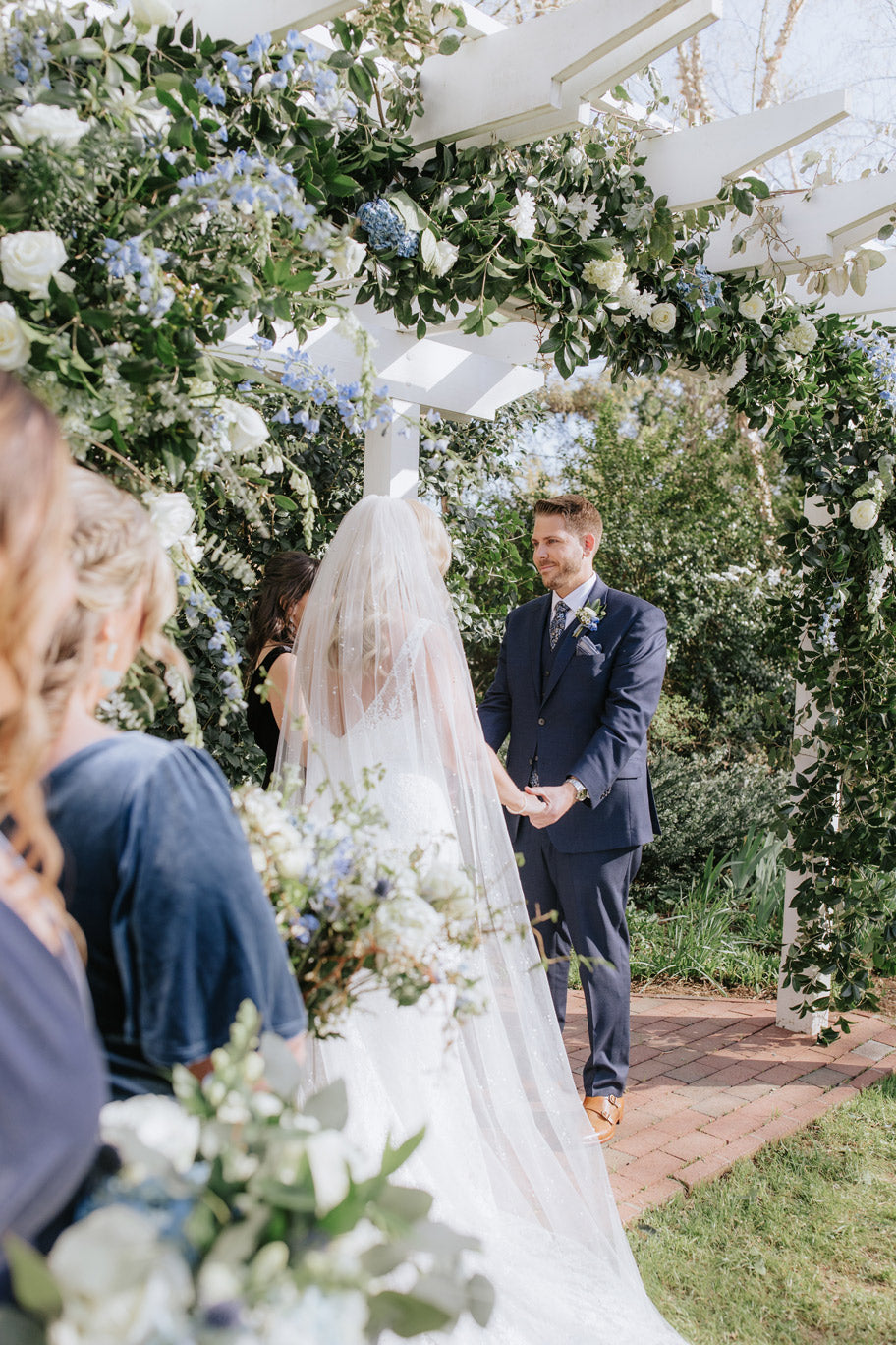 A bride and groom stand under a flower-adorned white arbor, holding hands and facing each other during their outdoor wedding ceremony, with bridesmaids in navy dresses in the foreground.