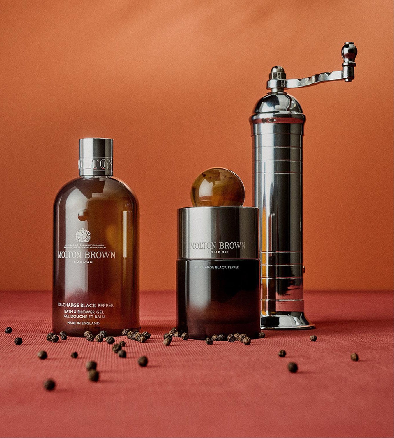 Three items on a red surface: a Molton Brown bath and shower gel, a Molton Brown eau de toilette, and a silver pepper grinder, with loose peppercorns scattered around. The background is a matching reddish shade.