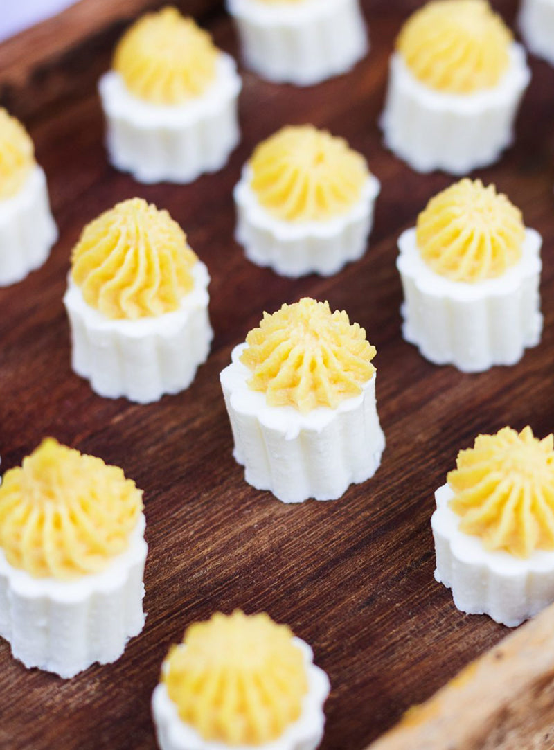 Small, white, cylindrical desserts with yellow piped tops are arranged neatly on a wooden tray. The treats have a ridged texture on the sides and swirled tops, resembling frosted pastries or confections.