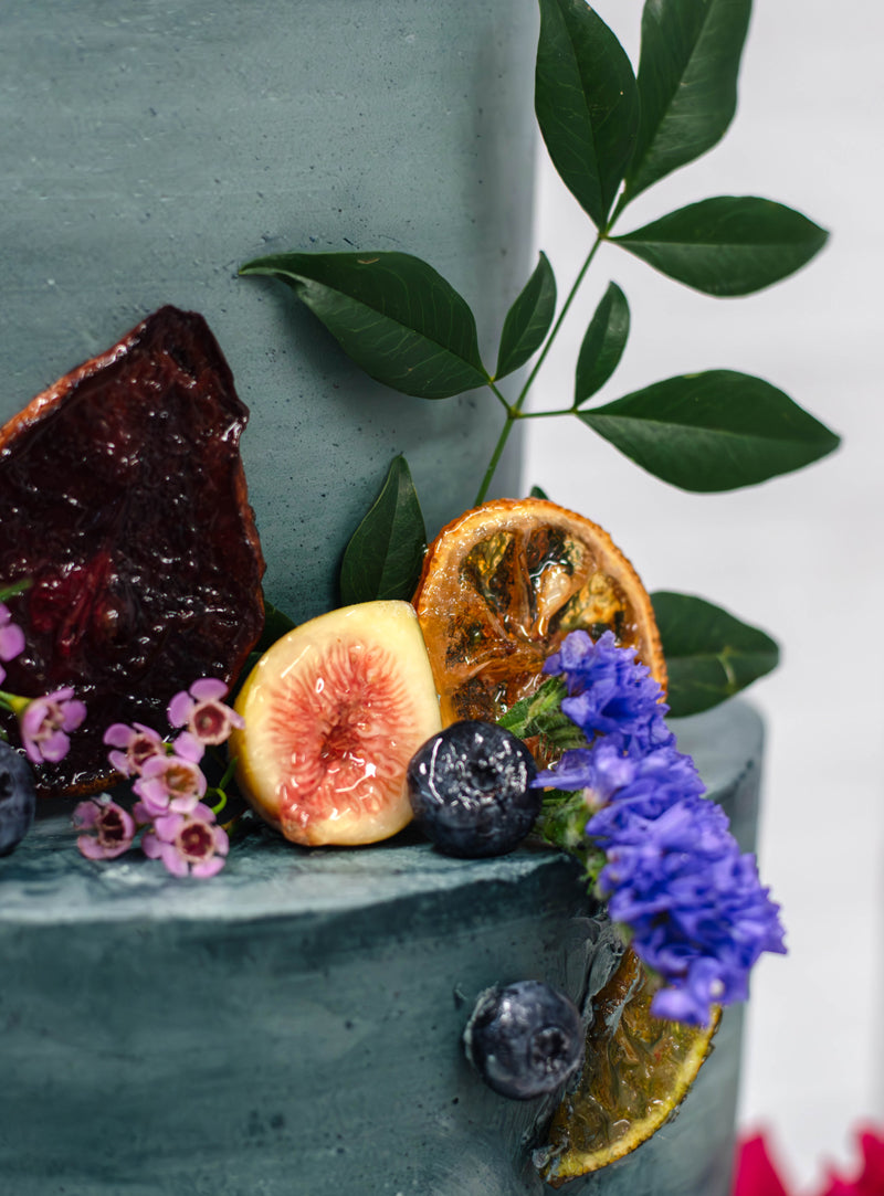A close-up of a dark green frosted cake decorated with fresh figs, dried citrus slices, blueberries, purple and pink flowers, and green leaves.
