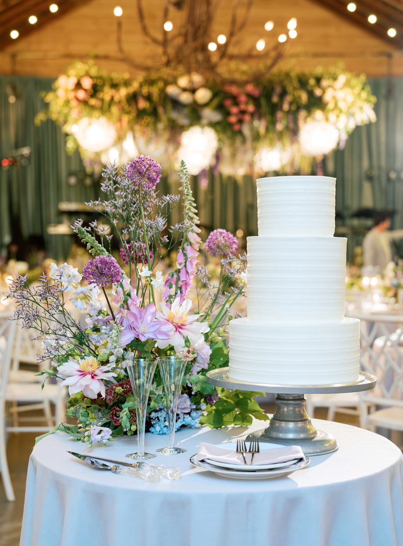 A white, three-tiered wedding cake sits on a round table next to a large floral arrangement with purple and pink flowers. The table is set with plates and utensils. Elegant decor and greenery are visible in the background.
