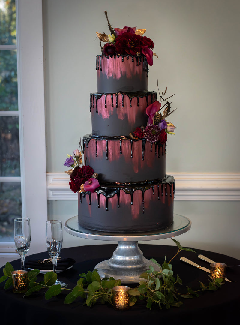 A four-tiered cake with black and metallic pink drip design, decorated with dark red and pink flowers, sits on a glass stand. The display includes candles, greenery, and two champagne flutes on a black tablecloth.