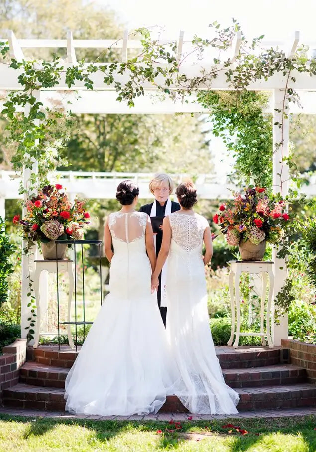 Two brides in white dresses stand hand in hand at an outdoor wedding ceremony under a floral arch, facing an officiant. Lush greenery and colorful flower arrangements decorate the scene.