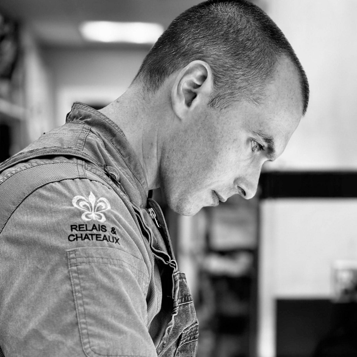 Black and white photo of a man in profile, focused intently. He wears a shirt with a "Relais & Chateaux" logo on the sleeve. The background is blurred, suggesting an indoor setting, possibly a kitchen.