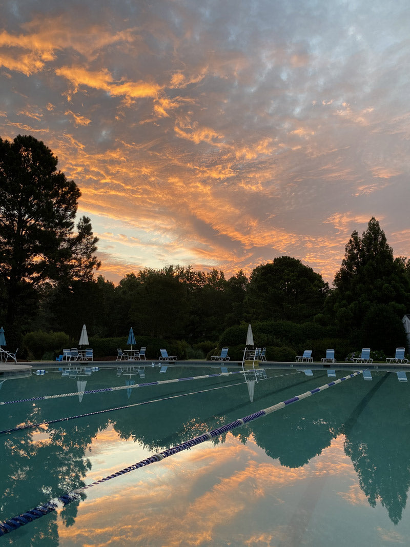 A calm outdoor swimming pool with lane dividers reflects a vibrant orange and blue sunset sky, surrounded by trees and lounge chairs with closed umbrellas.