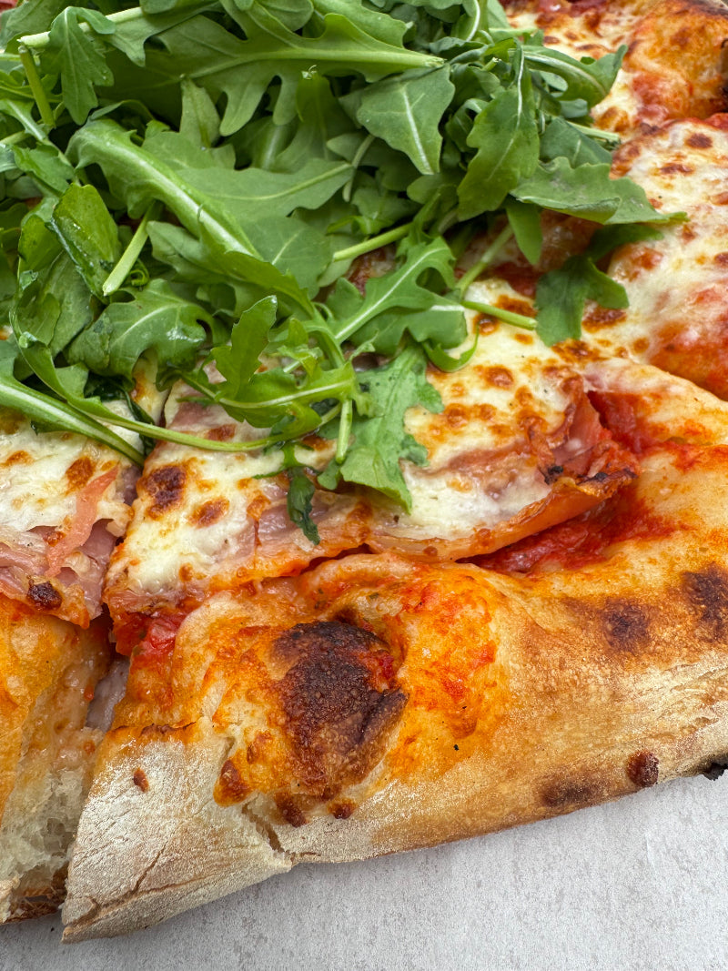 Close-up of a pizza topped with melted cheese, tomato sauce, ham, and a generous handful of fresh arugula on top of several slices. The crust is golden brown and slightly charred in spots.
