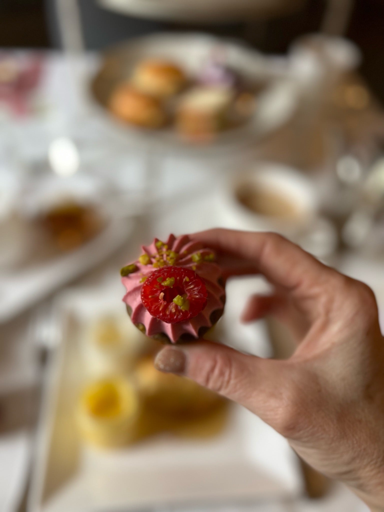 A hand holds a small, pink-frosted pastry topped with a raspberry and crushed pistachios. The background shows a table set with plates of assorted desserts and blurred cups.