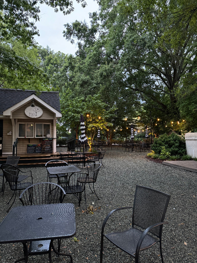 Outdoor café with empty black metal tables and chairs on a gravel patio, surrounded by trees and string lights. A small building with a menu sign is in the background under a cloudy sky.