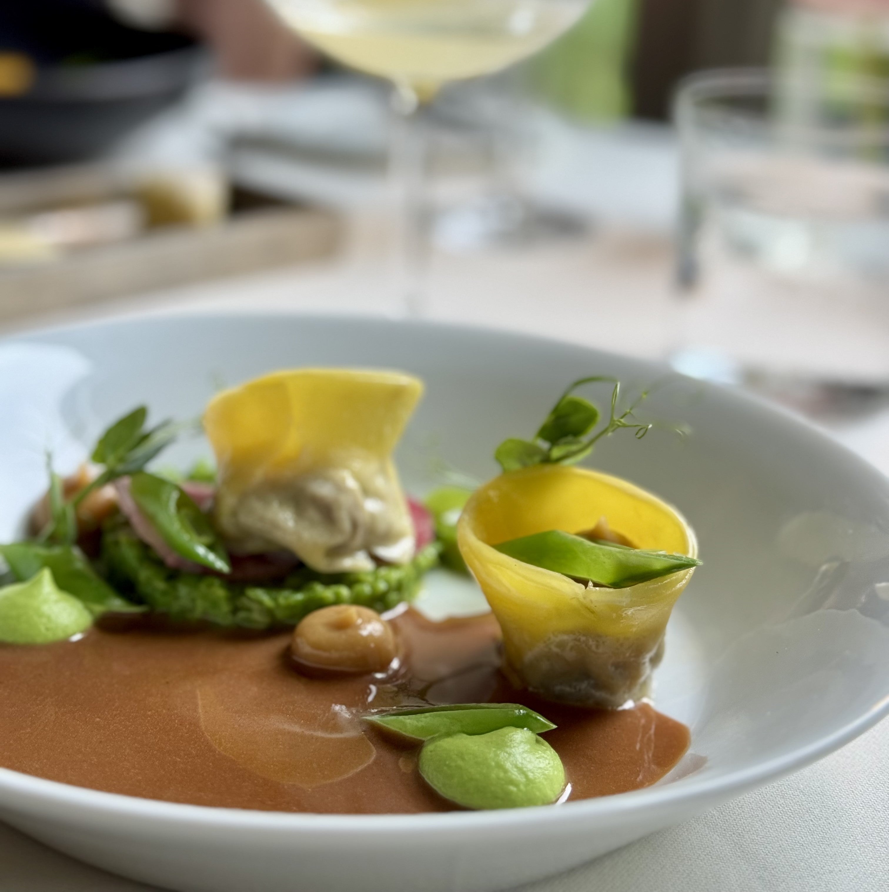 A gourmet dish featuring two delicate yellow pasta parcels, green pea purée, fresh greens, mushrooms, and rich brown sauce, artfully plated on a white dish with wine and water glasses in the background.