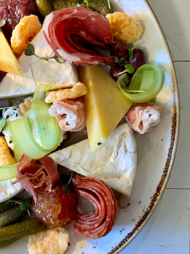 A close-up of a charcuterie platter featuring slices of cured meats, wedges of brie and hard cheese, pickles, green vegetable ribbons, small olives, and crispy cheese snacks on a white plate with a brown rim.