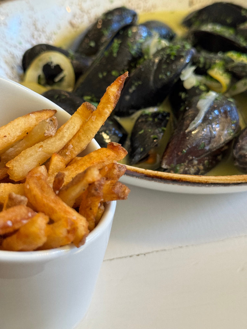 A close-up of a bowl of crispy French fries next to a plate of mussels in a creamy herb sauce. The dishes are served on a white table.