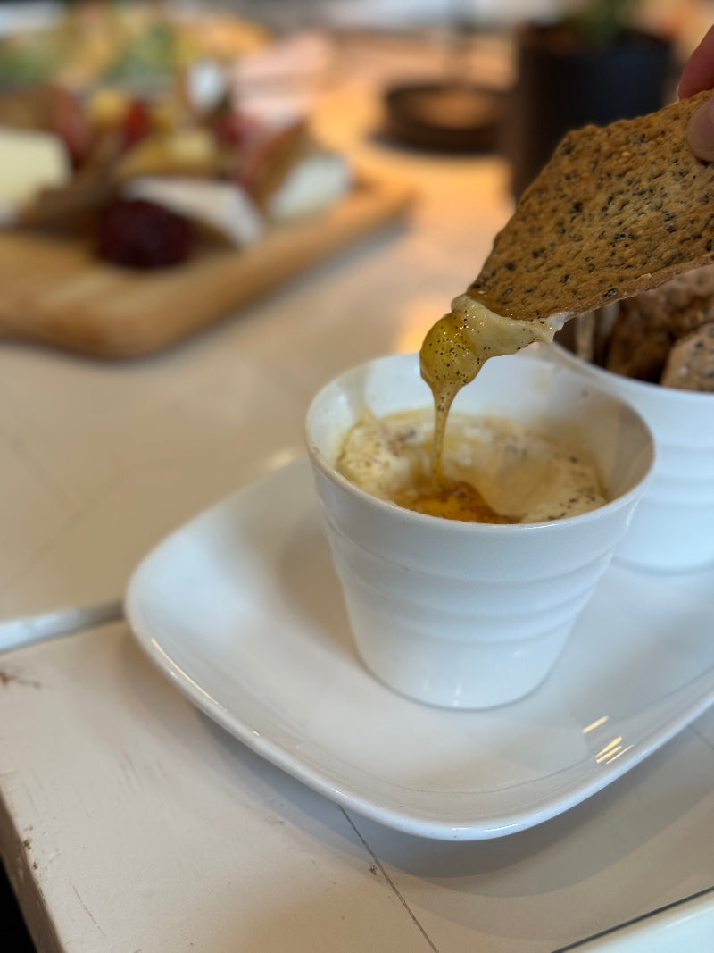 A hand dips a seeded cracker into a creamy dip in a white cup, with the dip stretching between the cracker and cup. A wooden board with assorted cheeses and snacks is blurred in the background.