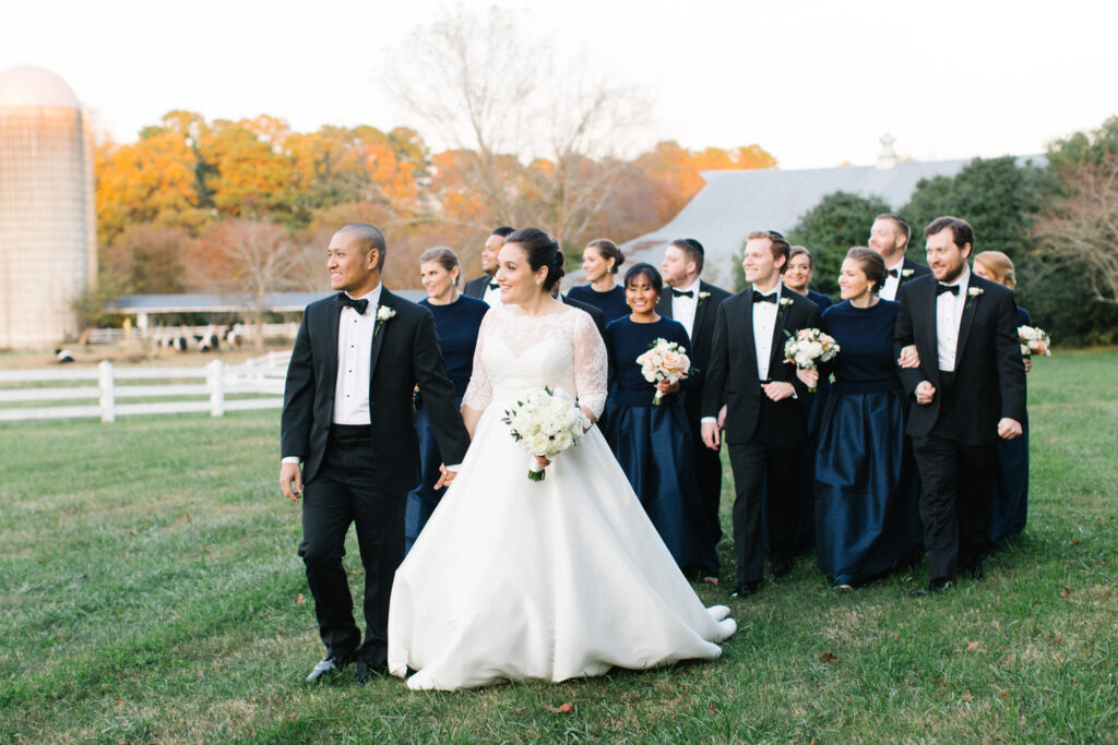 A bride and groom walk outside on grass with their wedding party, all dressed formally. The bridesmaids wear navy dresses, and the groomsmen wear black tuxedos. Autumn trees and a barn are visible in the background.