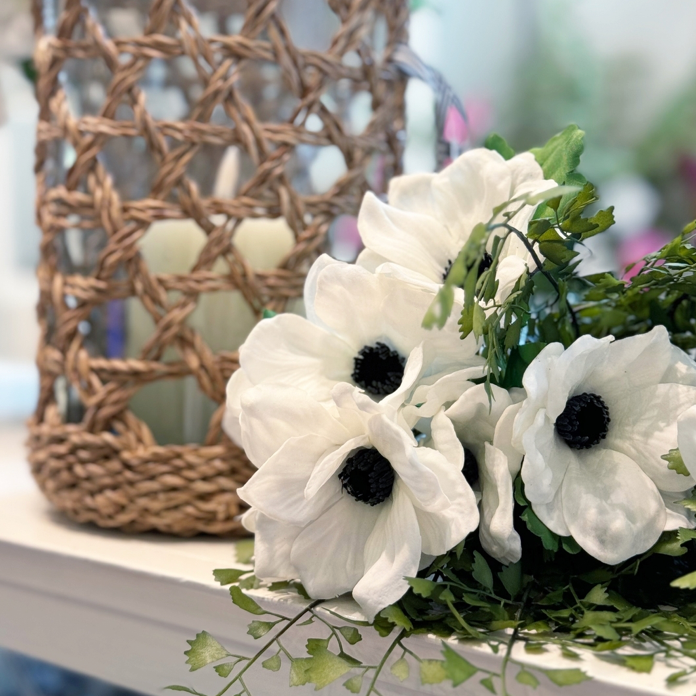 A close-up of white flowers with dark centers and green leaves arranged on a table next to a woven wicker lantern holding a candle. The background is softly blurred.
