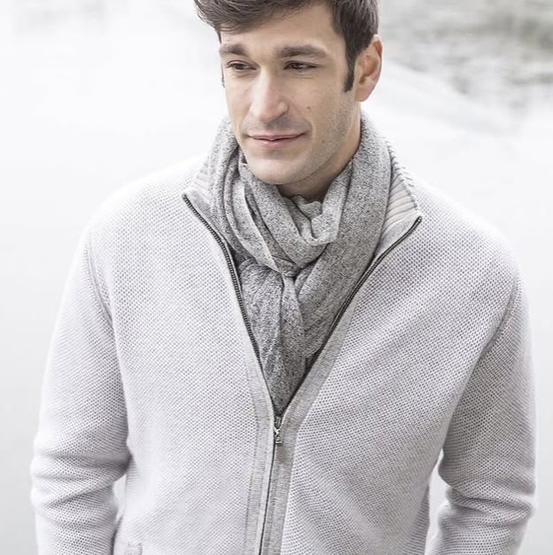 A man wearing a light grey textured zip-up sweater and a matching scarf stands outdoors near a body of water, looking off to the side with a neutral expression.