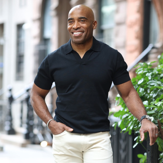 A smiling man wearing a black short-sleeve shirt and light-colored pants stands outdoors on a city sidewalk, leaning on a black railing next to green plants. The background features blurred buildings and steps.